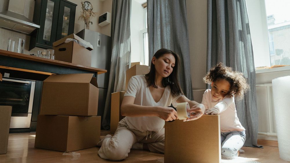 Kind sitzt zwischen Umzugskartons in einem noch leeren Zimmer und packt eine Kiste aus